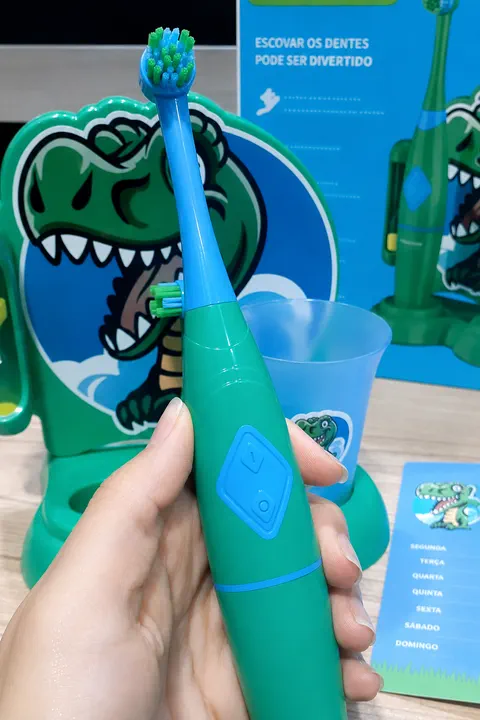 Escova elétrica verde segurada na mão, com a base do kit, o copo e a caixa do produto ao fundo