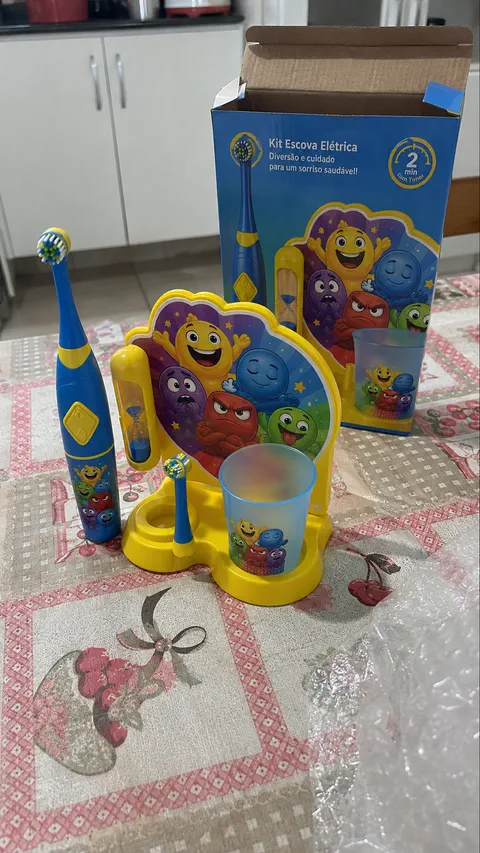 Kit amarelo montado sobre a mesa, com escova, ampulheta e copo, e a caixa do produto aberta atrás