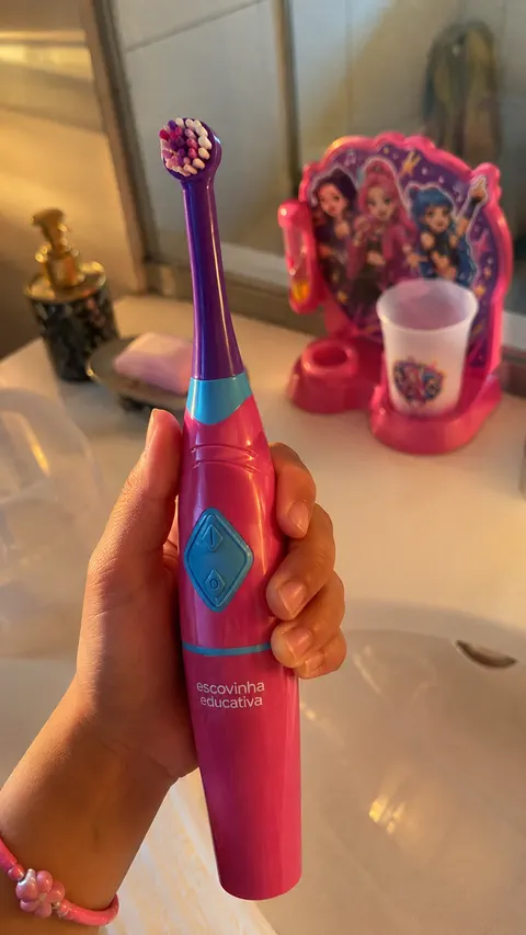 Escova elétrica rosa segurada na mão, com a base do kit e o copo apoiados na pia ao fundo